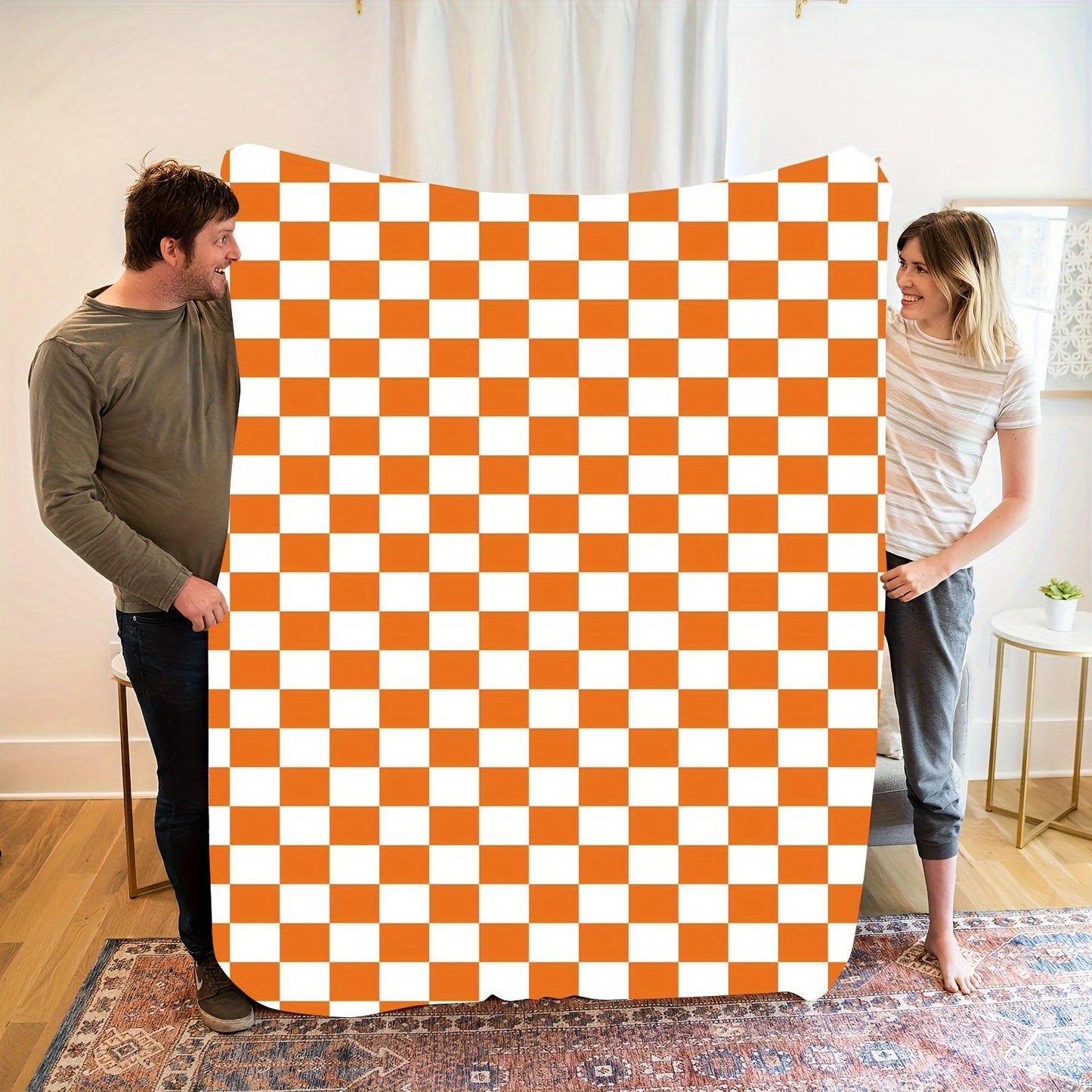 One piece of an orange and white checkered print fleece throw blanket featuring a geometric pattern. This all-season blanket is machine washable, durable, and anti-tear, making it perfect for use in the bedroom, living room, or while camping. It weighs