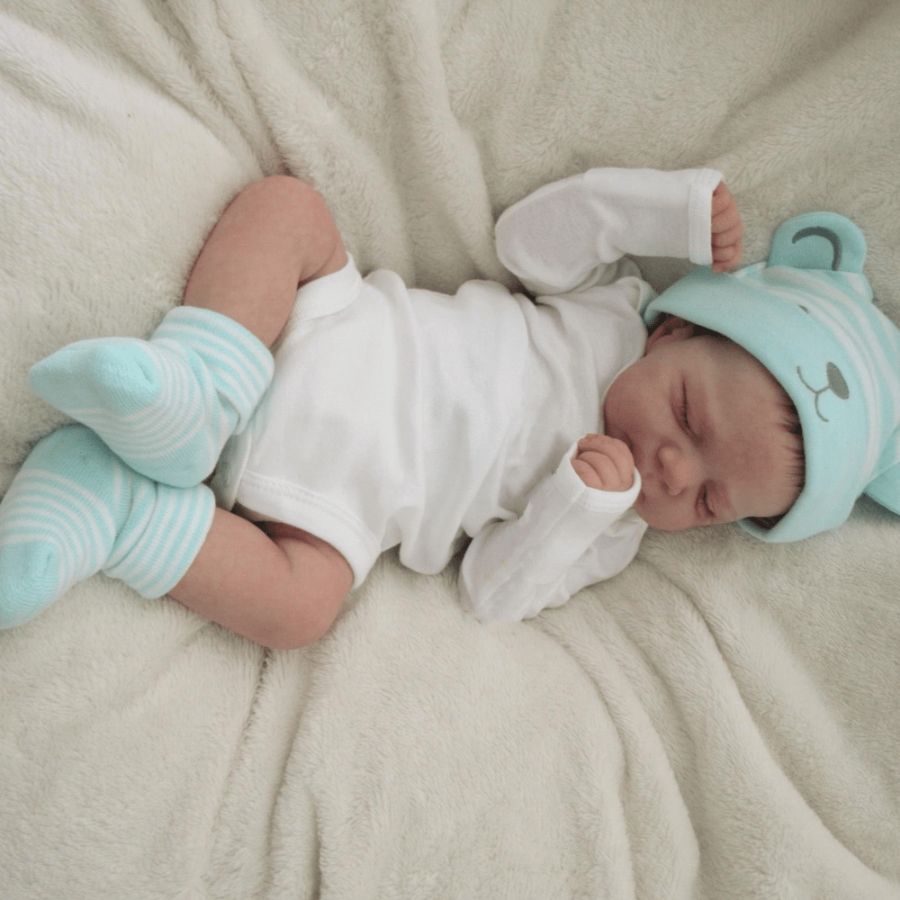 Realistic 18-inch reborn girl doll with blue bear hat, soft vinyl body, detailed skin, visible veins, and lifelike sleeping pose. Ideal birthday gift for children 3 and up.