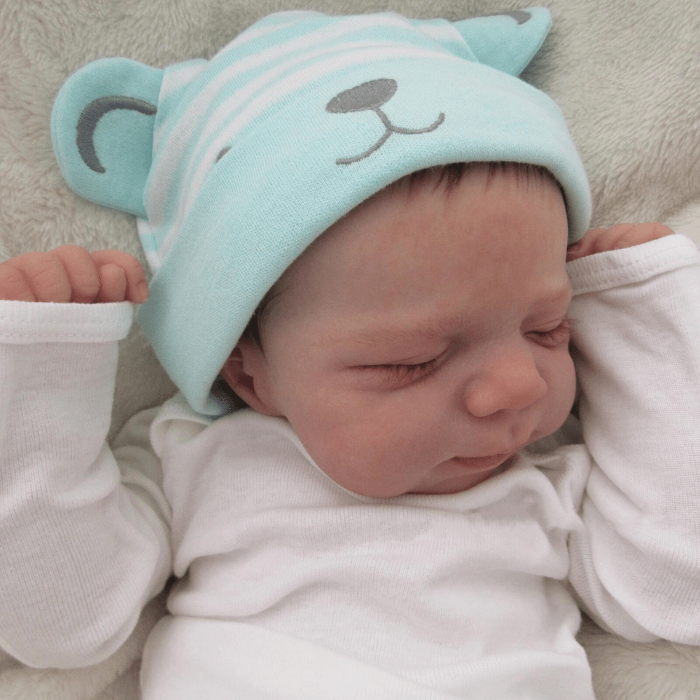 Realistic 18-inch reborn girl doll with blue bear hat, soft vinyl body, detailed skin, visible veins, and lifelike sleeping pose. Ideal birthday gift for children 3 and up.