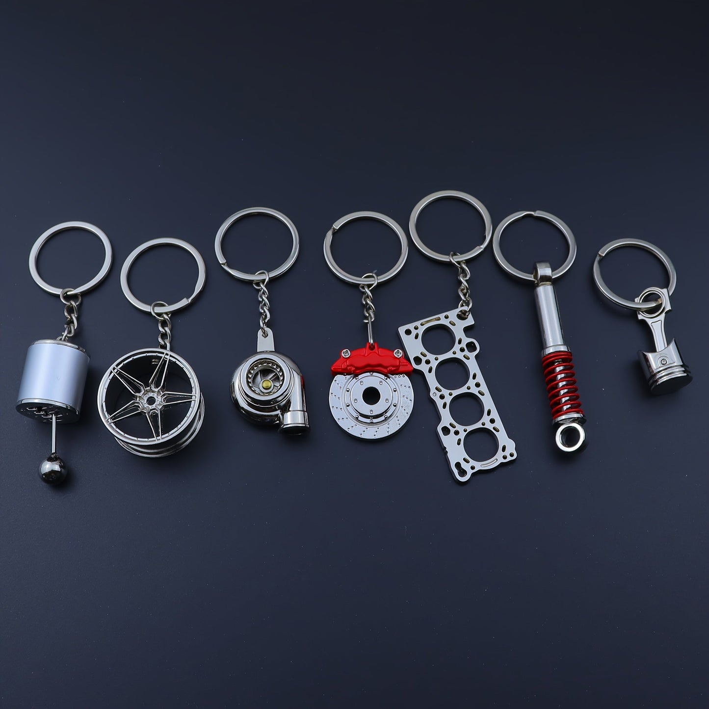 Funky car modification keychains set includes seven pieces: piston, turbo, wheel hub, gear shift, shock absorber, brake disc, and engine block. Made of zinc alloy in a sleek black finish.