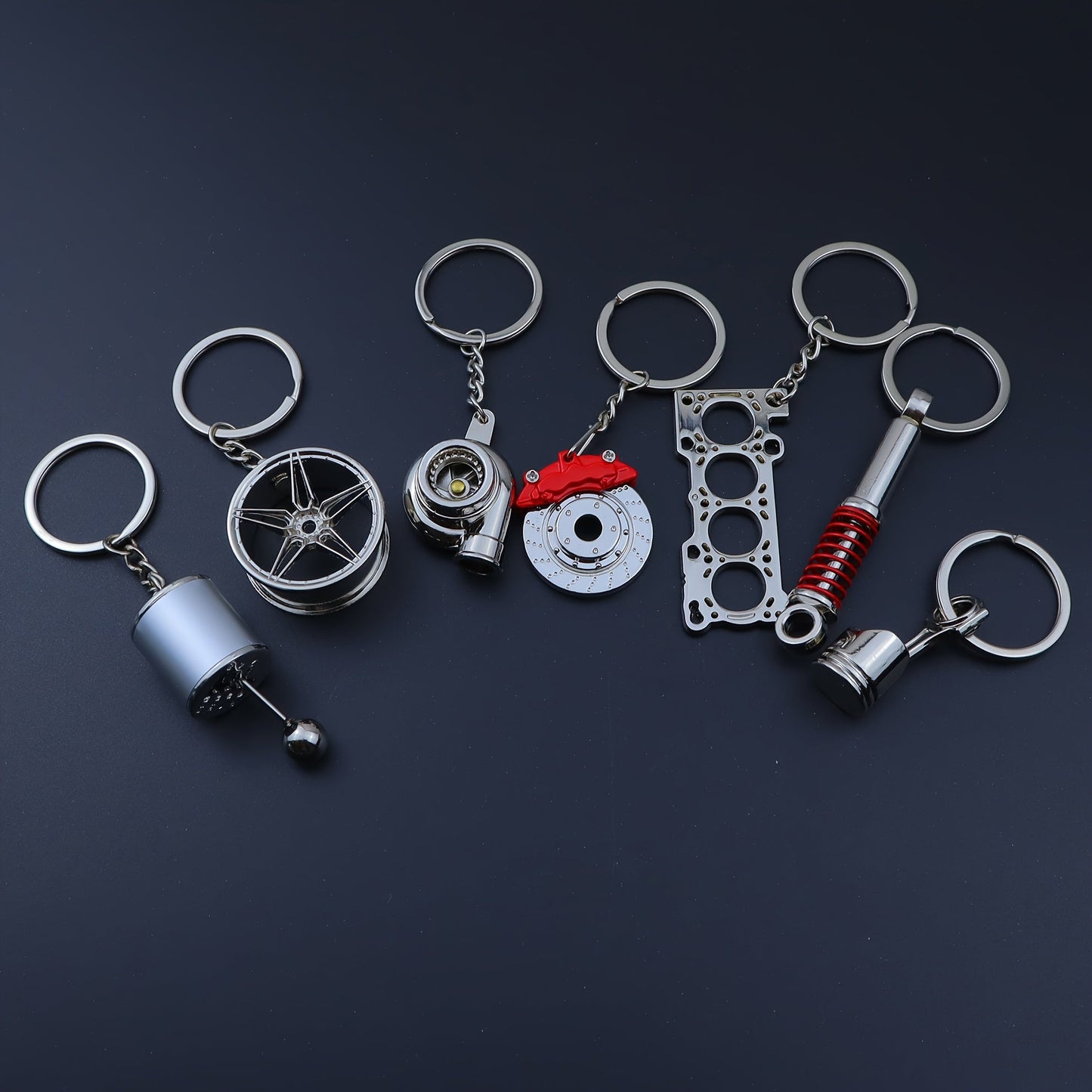 Funky car modification keychains set includes seven pieces: piston, turbo, wheel hub, gear shift, shock absorber, brake disc, and engine block. Made of zinc alloy in a sleek black finish.