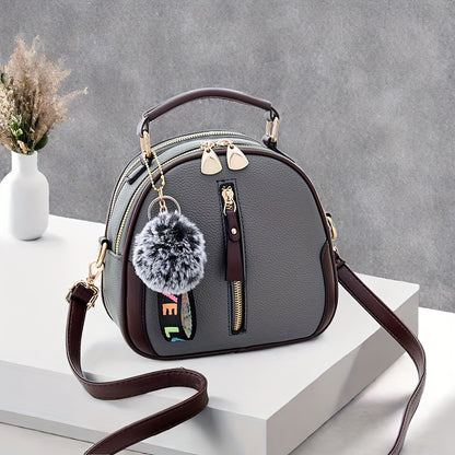 Large capacity women's handbag in brown and gray with golden-tone hardware, adjustable strap, and zipper closure. Features pom pom charm and casual yet trendy design.