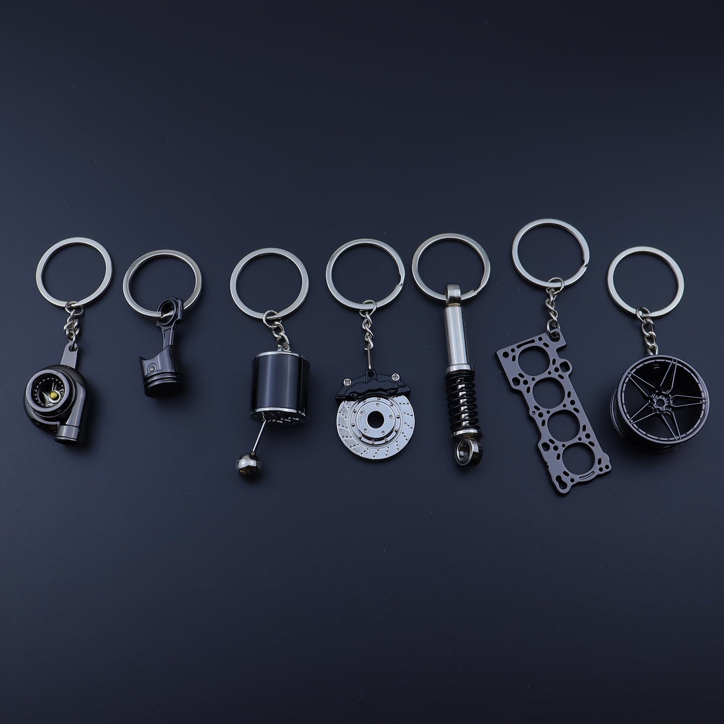 Funky car modification keychains set includes seven pieces: piston, turbo, wheel hub, gear shift, shock absorber, brake disc, and engine block. Made of zinc alloy in a sleek black finish.