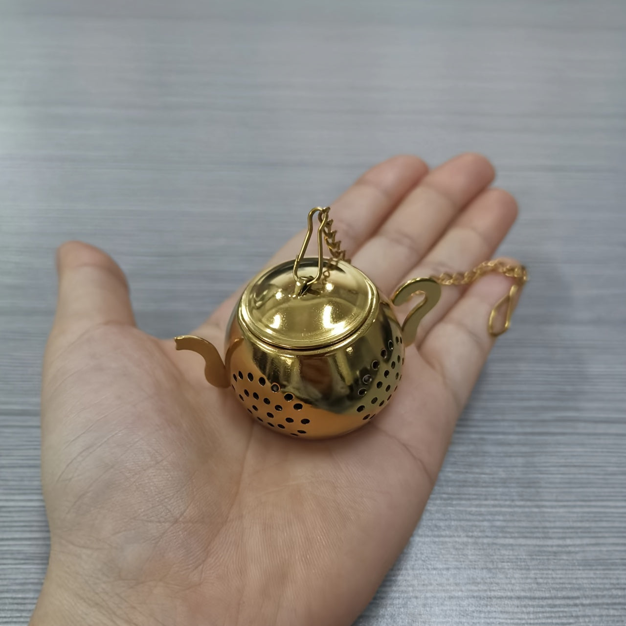 Golden rose stainless steel teapot-shaped tea strainer with uniform filter holes and hanging chain for cups.