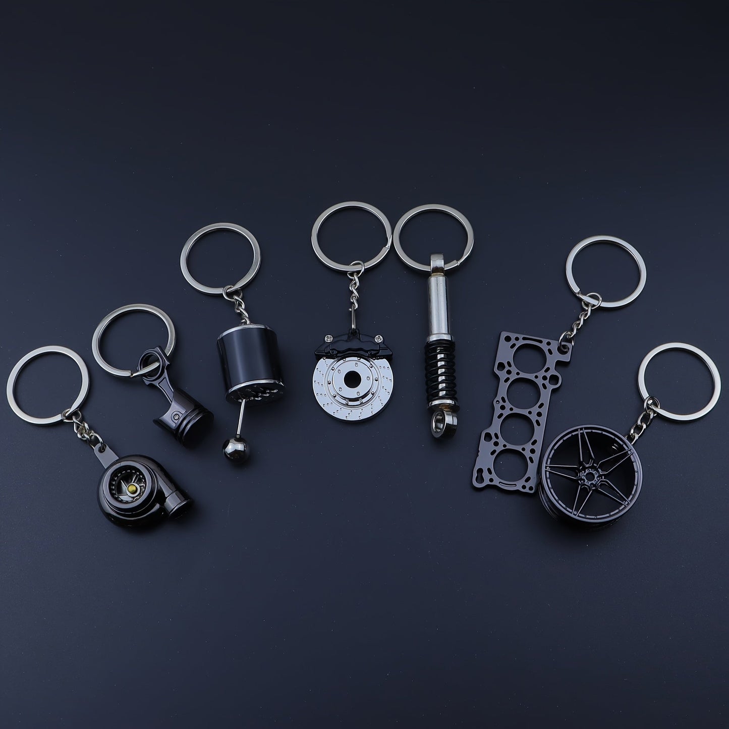 Funky car modification keychains set includes seven pieces: piston, turbo, wheel hub, gear shift, shock absorber, brake disc, and engine block. Made of zinc alloy in a sleek black finish.