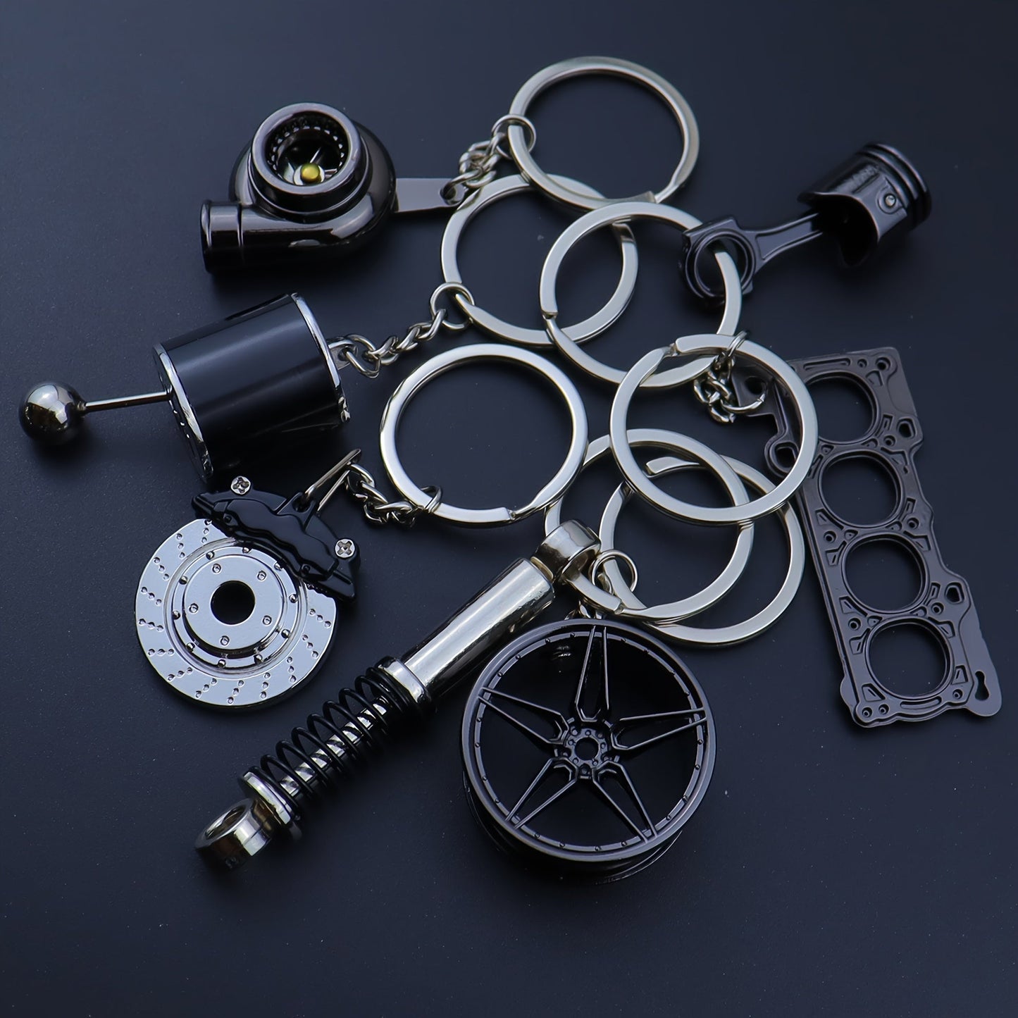 Funky car modification keychains set includes seven pieces: piston, turbo, wheel hub, gear shift, shock absorber, brake disc, and engine block. Made of zinc alloy in a sleek black finish.