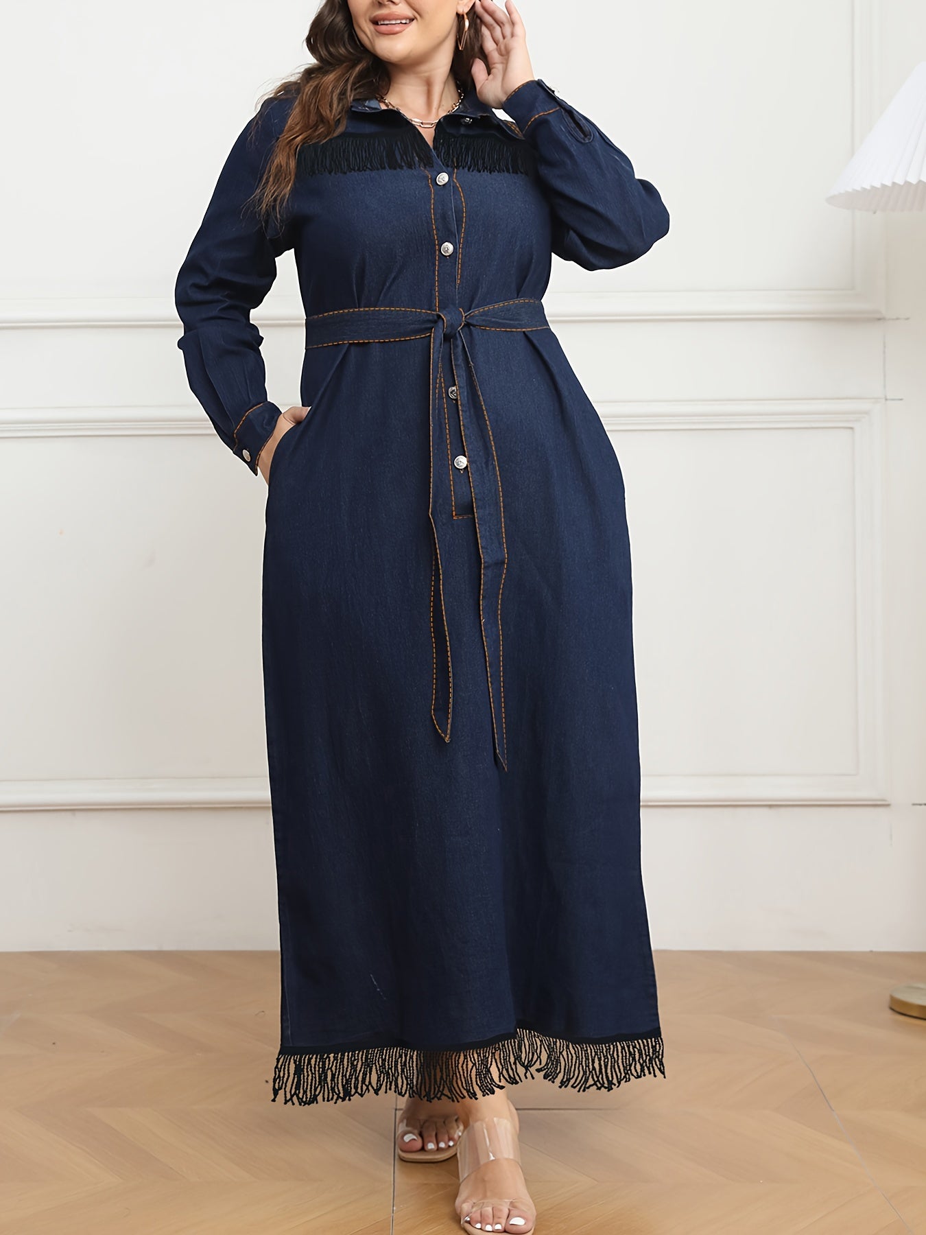 Women's plus size navy blue denim dress with long sleeves, button-up shirt collar, fringe hem, belt detail, and cotton/polyester blend. Machine washable.