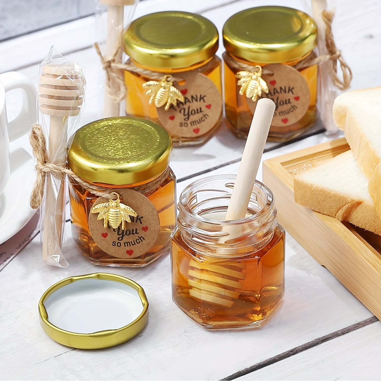 Small hexagonal glass honey jars come in 1.5 oz sizes in sets of 20, 30, 50, or 60. Each jar includes a spoon, bee symbol, and thank you card. They have golden lids and are ideal for baby gifts, weddings, and parties.