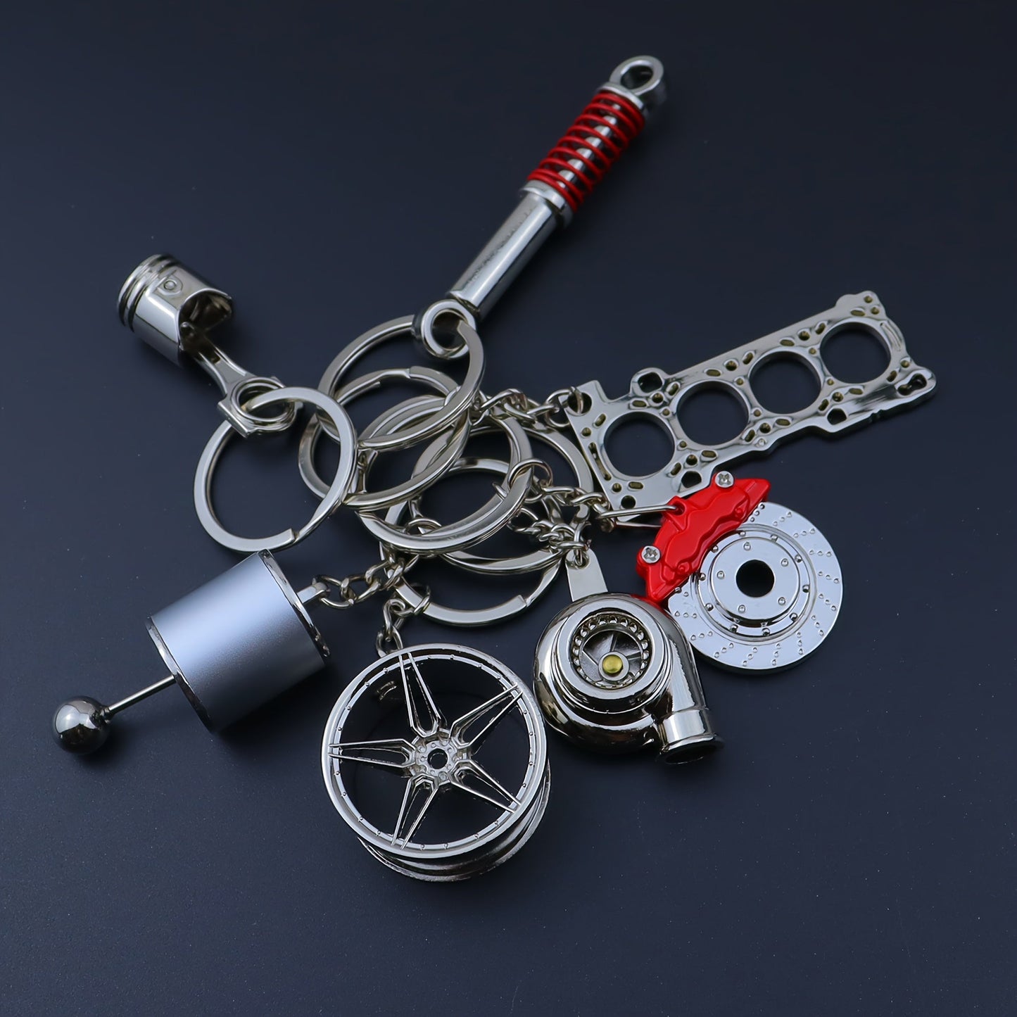 Funky car modification keychains set includes seven pieces: piston, turbo, wheel hub, gear shift, shock absorber, brake disc, and engine block. Made of zinc alloy in a sleek black finish.