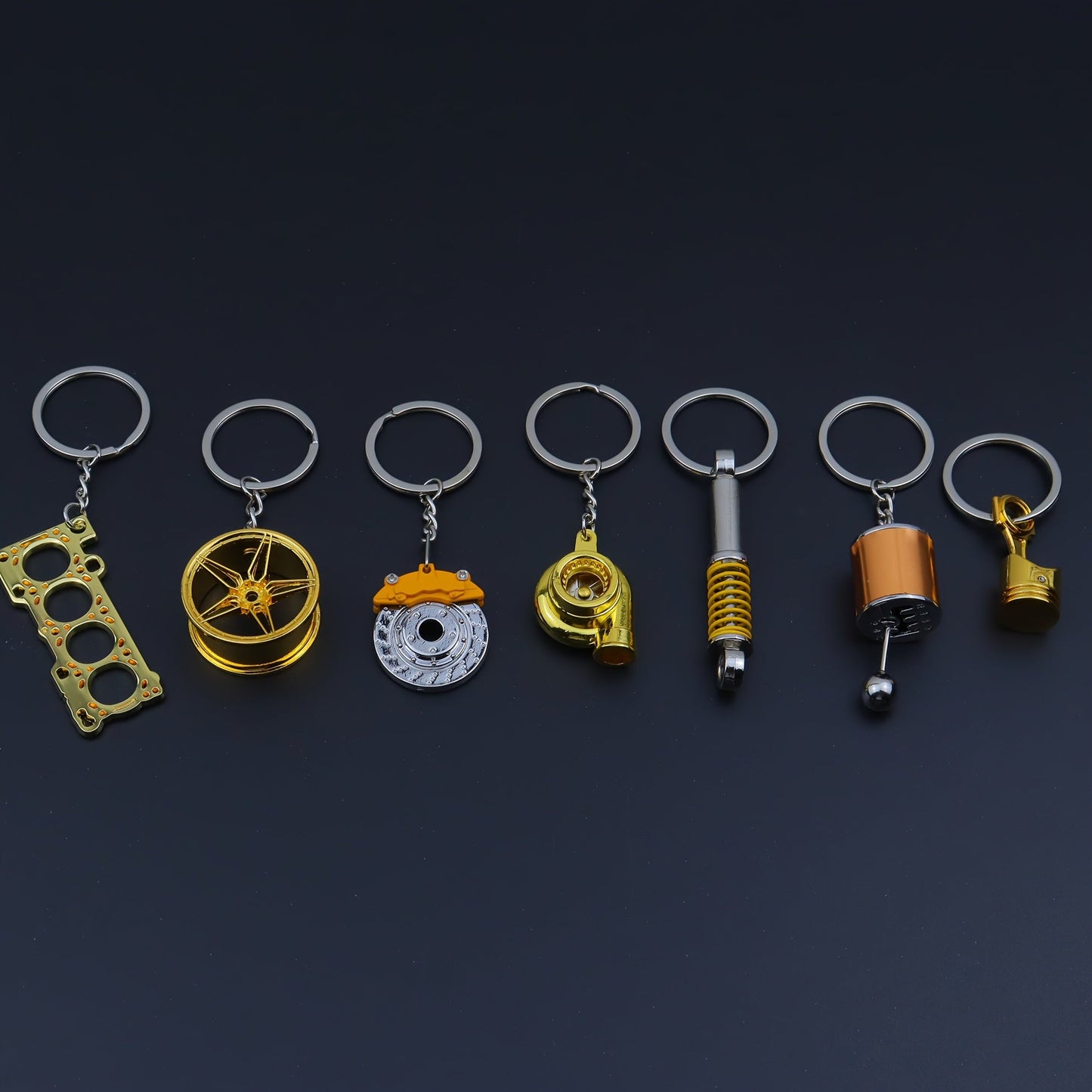 Funky car modification keychains set includes seven pieces: piston, turbo, wheel hub, gear shift, shock absorber, brake disc, and engine block. Made of zinc alloy in a sleek black finish.